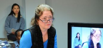 A woman wearing glasses looks forward with a serious expression; a TV screen beside her shows her and another seated woman in the background.