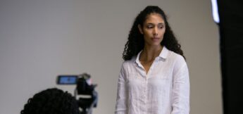 A woman with curly hair wearing a white button-up shirt stands in front of a camera and looks to the side against a plain background.