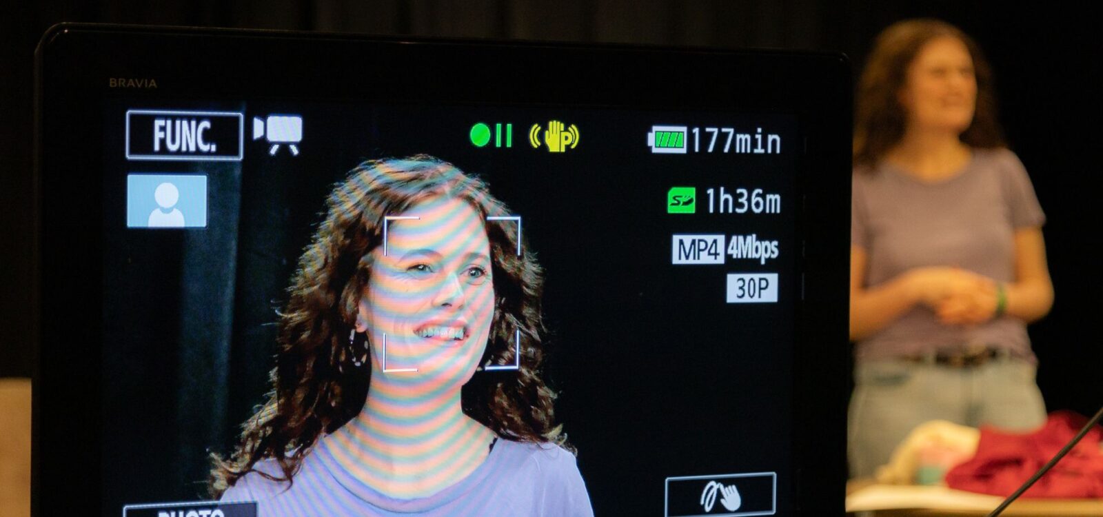 A woman stands in front of a table, her face framed and displayed on a video camera screen in the foreground, with recording indicators visible.