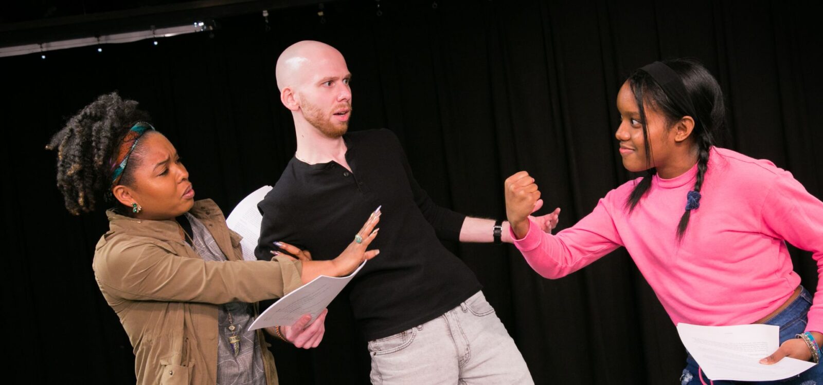 Three people hold scripts while acting; one person is being held back as they raise a fist, another looks surprised, and a third tries to keep peace. They are on a stage with a black curtain.