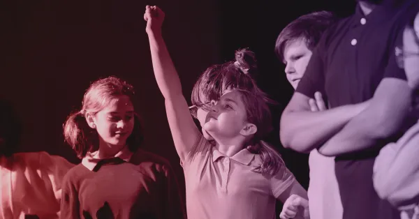 A group of children stand together; one girl in the center raises her arm enthusiastically while others look on.