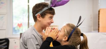 Two children wearing animal ear headbands face each other, with one whispering into the other's ear while both smile indoors.