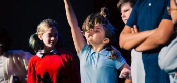 A group of children stand together, with one girl in a blue shirt raising her arm enthusiastically while others look on.