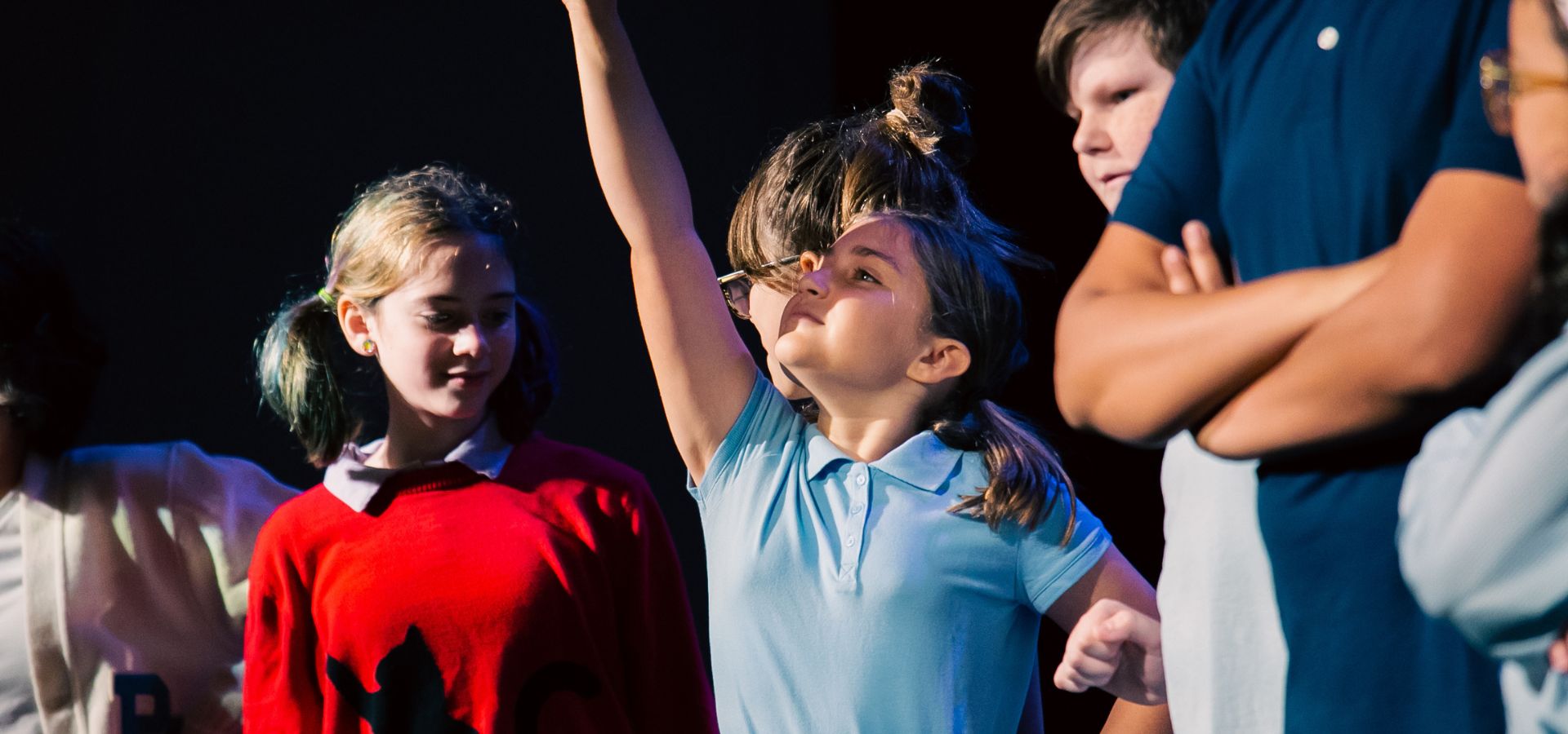 A group of children stand together, with one girl in a blue shirt raising her arm enthusiastically while others look on.