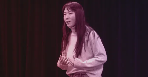 A woman with long hair and a light sweater stands in front of a dark curtain, gesturing with her hands as she speaks.