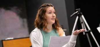 A woman with shoulder-length brown hair holds papers and gestures with her hand while speaking, seated indoors near a tripod.