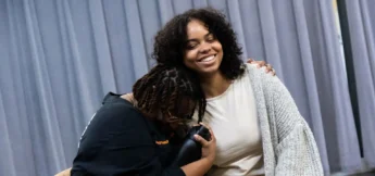 Two people sit together; one person is hugging and leaning on the other, who is smiling with an arm around them. A gray curtain hangs in the background.