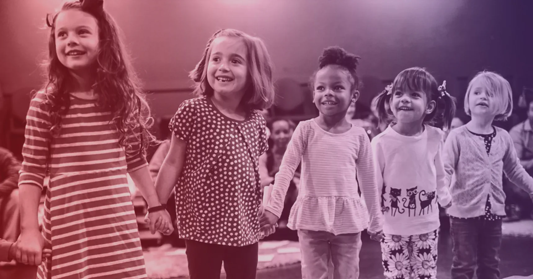 Five young children stand in a row, holding hands and smiling, with other children seated in the blurred background.