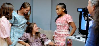 Four girls in vintage-style dresses talk and smile while one sits on a chair; another woman operates a video camera nearby.