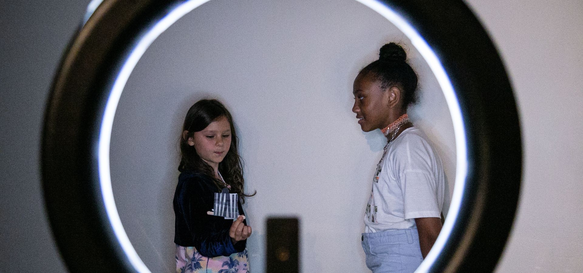 Two girls stand against a white wall, framed by a circular ring light; one holds a smartphone while the other looks at her.