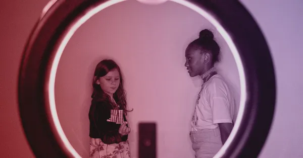 Two girls stand against a wall, viewed through a circular frame with a glowing ring of light. One girl looks down, the other looks at her.