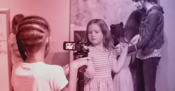A child films other children with a video camera on a tripod while two girls and an adult interact in the background.