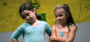 Two young girls stand side by side in front of a yellow background, both with arms crossed, looking to the side with neutral or slightly pensive expressions.
