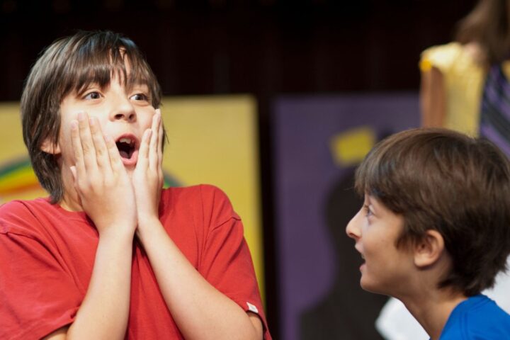 Two children are interacting, with one boy in a red shirt holding his face in apparent surprise, while another child looks at him. A girl stands blurred in the background.