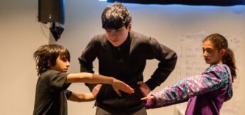 Three children stand together inside; one stands with hands on hips while the other two extend their arms forward in front of them.