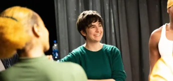 A person in a green shirt smiles while standing with others in a room with dark curtains in a free improv comedy workshop.