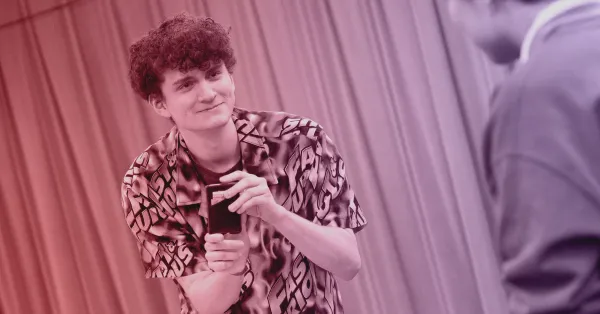 A young man with curly hair holds a smartphone and smiles at another person indoors, with curtain panels in the background.