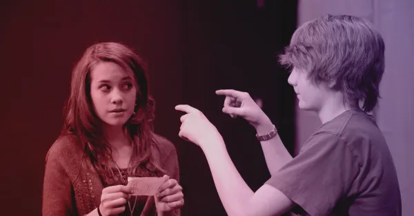 Free Improv612 4 Two teenagers stand indoors; one holds a piece of paper and looks to the side, while the other gestures with both hands, pointing forward.