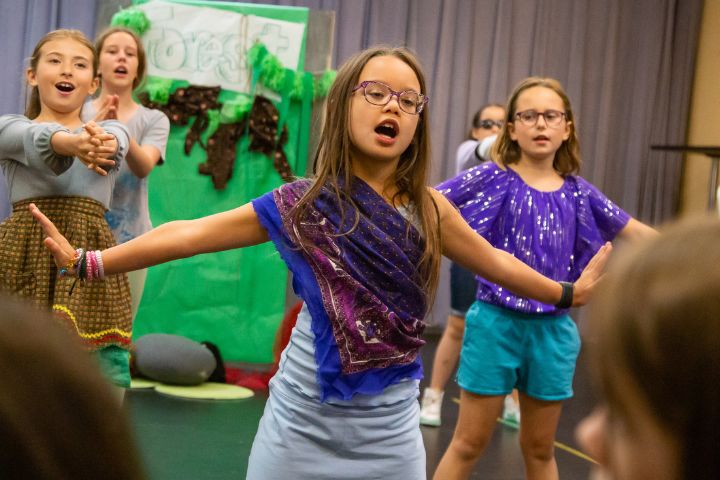 Children perform on stage in colorful costumes with arms outstretched, in front of a green scenery backdrop and gray curtain.