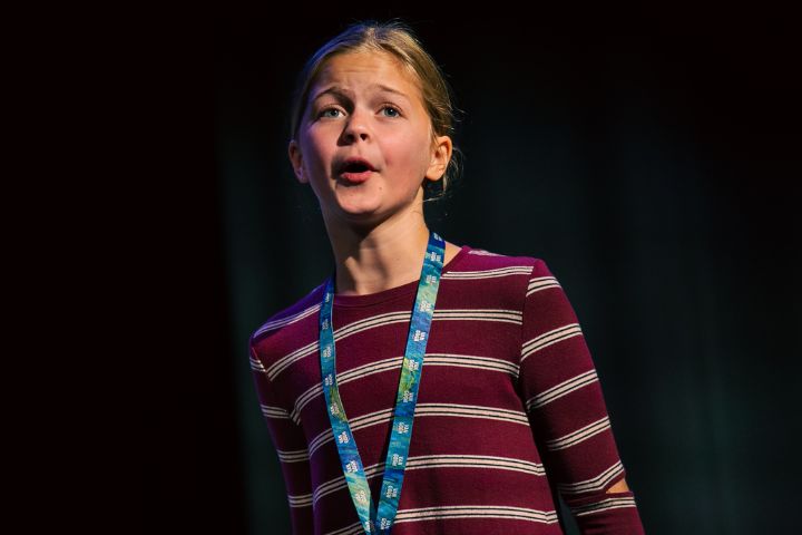 A girl with a blue lanyard and striped shirt speaks or sings on stage against a dark background.