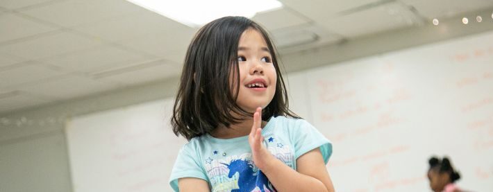 A young girl in a blue shirt stands in a classroom, smiling and clapping her hands, with two other children blurred in the background.