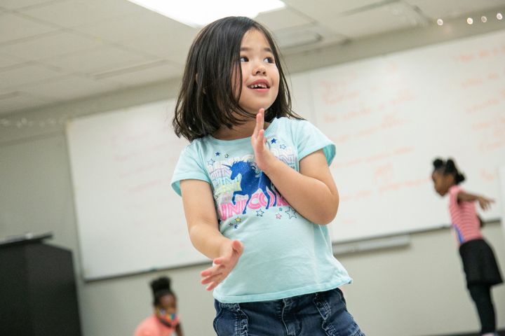 Free Mt Wksp Gk 2 A young girl in a blue shirt stands in a classroom, smiling and clapping her hands, with two other children blurred in the background.
