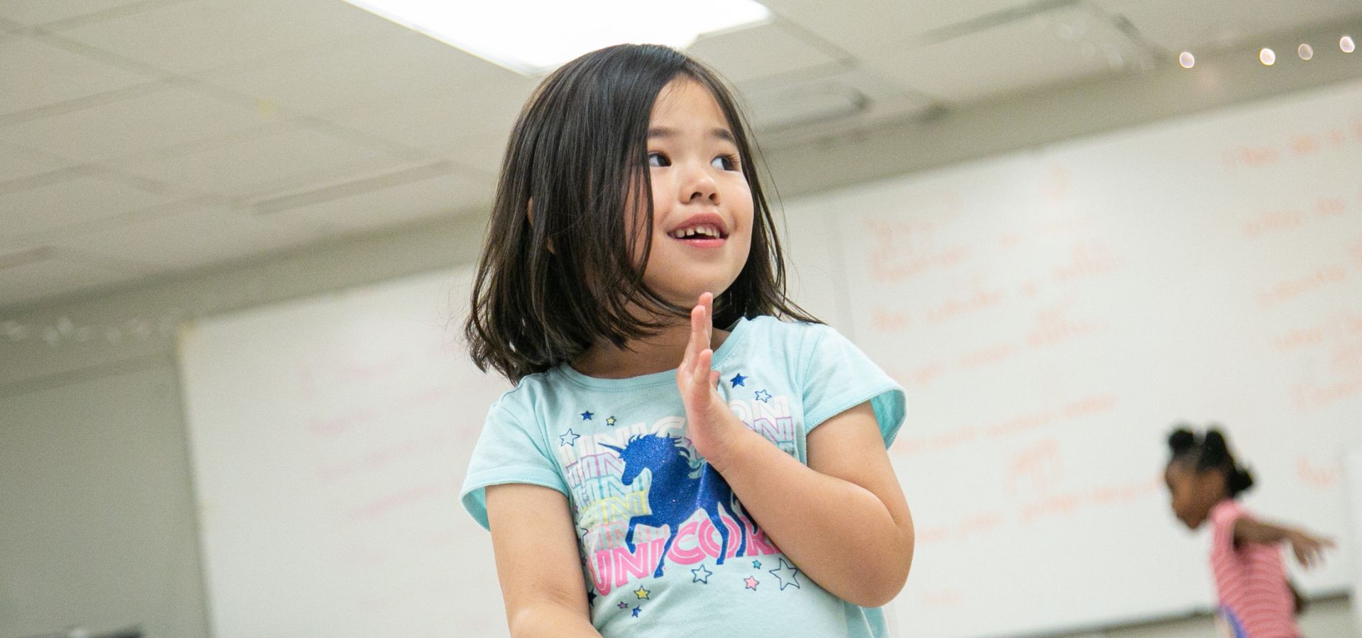 A young girl with medium-length dark hair and a light blue unicorn shirt stands indoors, smiling and looking to the side. A whiteboard and another child are visible in the background.