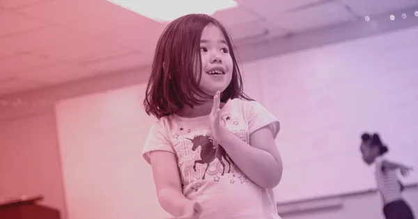 A young girl stands indoors, smiling with her hands together. A blurred child is visible in the background.