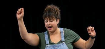 A person with curly hair wearing a green shirt and denim overalls raises both arms while speaking, against a black background.