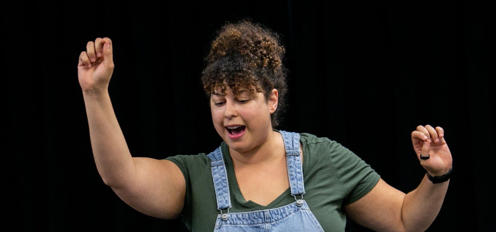 A person with curly hair wearing a green shirt and denim overalls raises both arms while speaking, against a black background.