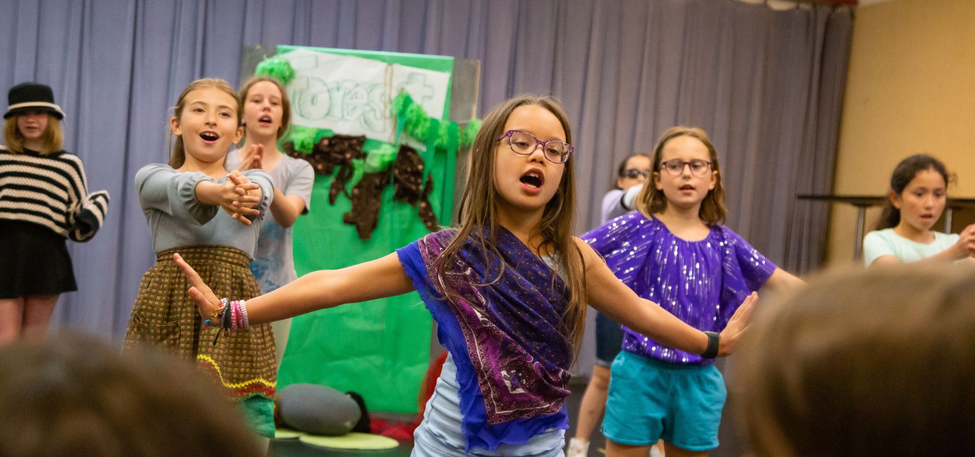 A group of children perform on stage, with one girl in the center extending her arms and singing while others stand and gesture behind her, in front of a green forest backdrop.