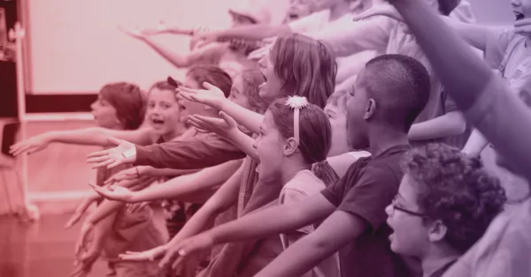 A group of children stand in rows with arms outstretched, appearing to sing or perform together in a classroom or rehearsal setting.