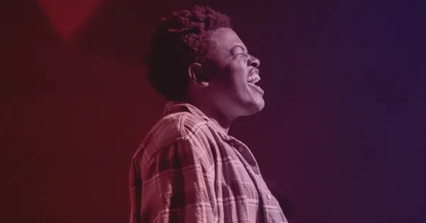 Person in a plaid shirt stands in profile against a red and purple background, appearing to sing or speak passionately.