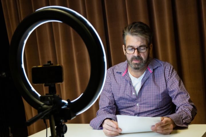 A man with glasses sits at a table reading from a paper, with a smartphone and ring light set up in front of him, against a brown curtain background.