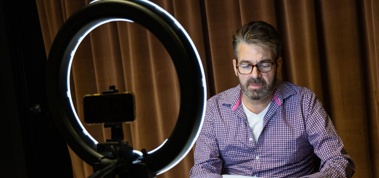 A man with glasses sits in front of a smartphone and ring light, reading or working, with a brown curtain in the background.