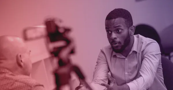 A man in a button-down shirt speaks to another person across a table, with a video camera recording their conversation in the foreground.