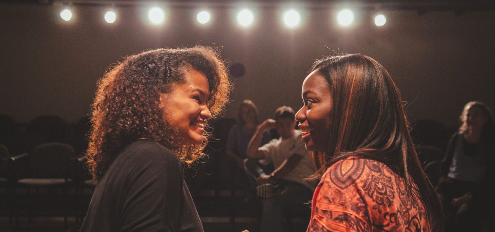 Two women stand face to face having a conversation in a dimly lit room with bright lights overhead and people seated in the background.
