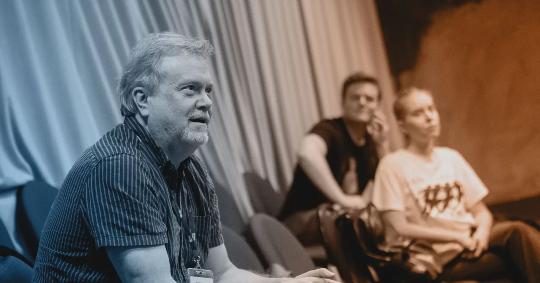 A man with a beard sits in the foreground, speaking or listening, while two people sit in the background, attentively watching. The setting appears to be a discussion or seminar.