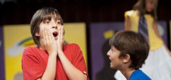 A boy in an improv comedy class in a red shirt looks surprised with his hands on his cheeks, while another boy faces him and a girl stands in the background.