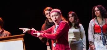 A high school actor in a red tracksuit and headband gestures animatedly on stage at improv camp, with three other people standing behind her, all under stage lighting.