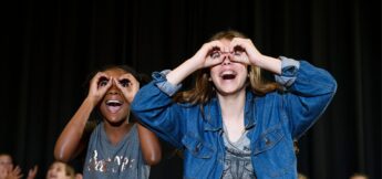 Two children stand in front of a black background, smiling and making pretend binoculars with their hands held up to their eyes in improv camp.