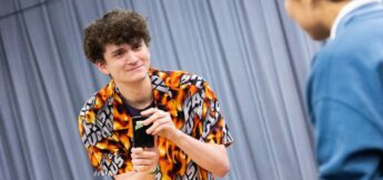 A young person in a flame-patterned shirt holds a black object and smiles while interacting with someone in a blue top indoors.