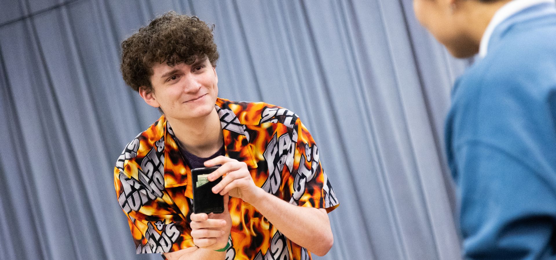 A young person in a flame-patterned shirt holds a black object and smiles while interacting with someone in a blue top indoors.