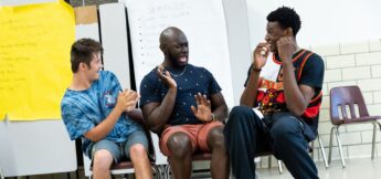 Three young men sit on chairs indoors, facing each other and laughing, with whiteboards and yellow paper visible in the background.