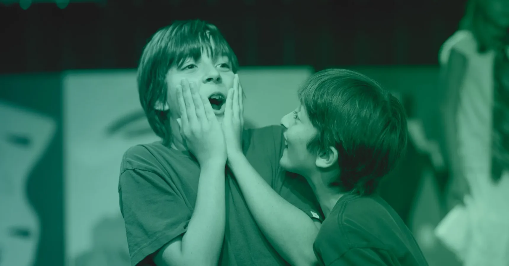 Two children face each other; one child holds their own cheeks with a surprised expression while the other smiles at them and touches their arm.