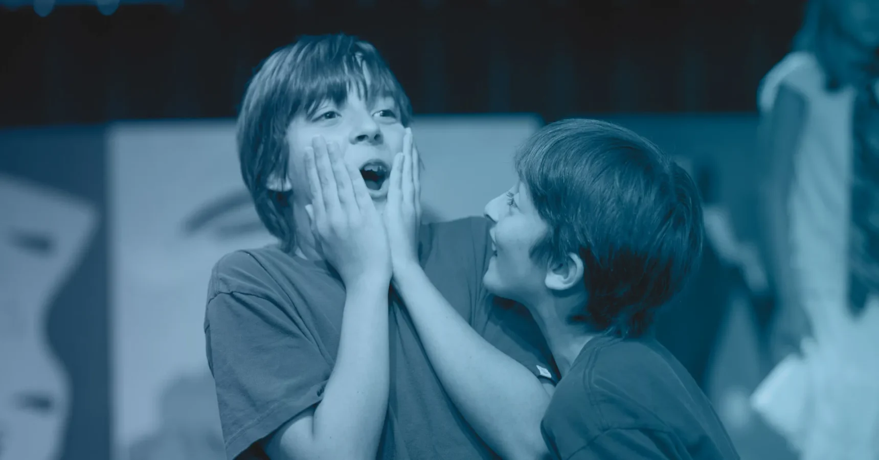 Two children wearing t-shirts interact; one child holds their face in surprise while the other smiles and reaches toward them. The image has a blue color filter.