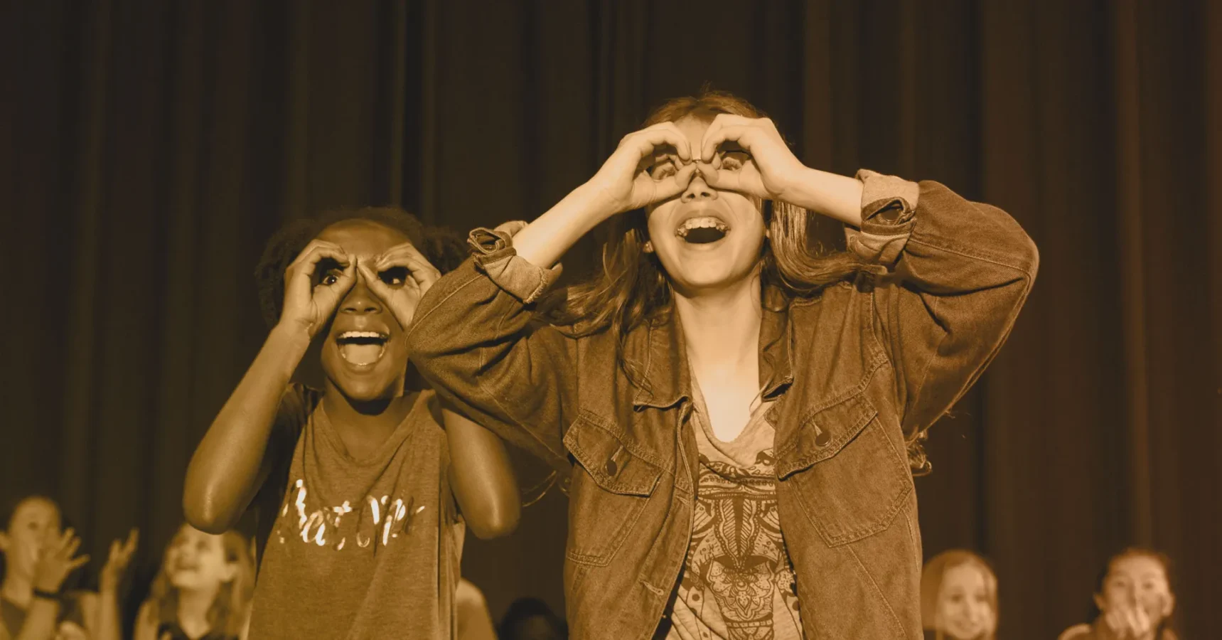 Two children on stage smile and make pretend binoculars with their hands in front of their eyes; others are visible in the background.