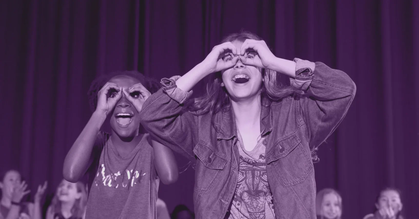 Two children stand in front of a dark curtain, smiling and making binocular shapes with their hands over their eyes.