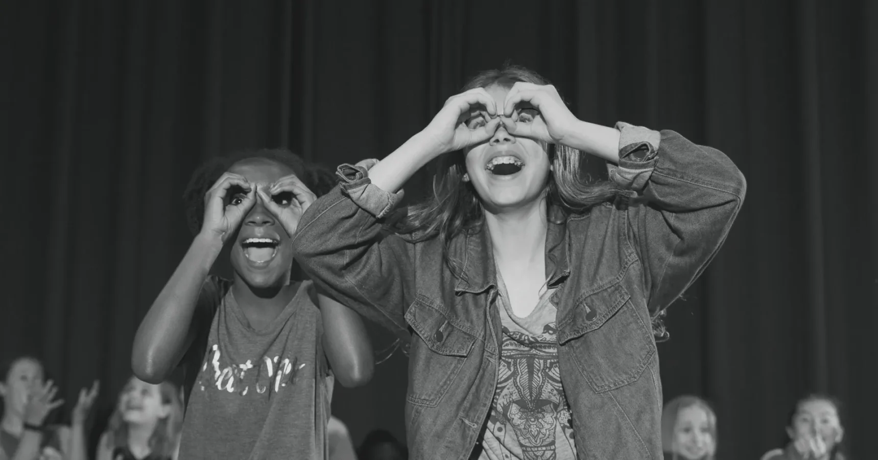Two children stand side by side, smiling and making binocular shapes with their hands over their eyes in front of a dark curtain.
