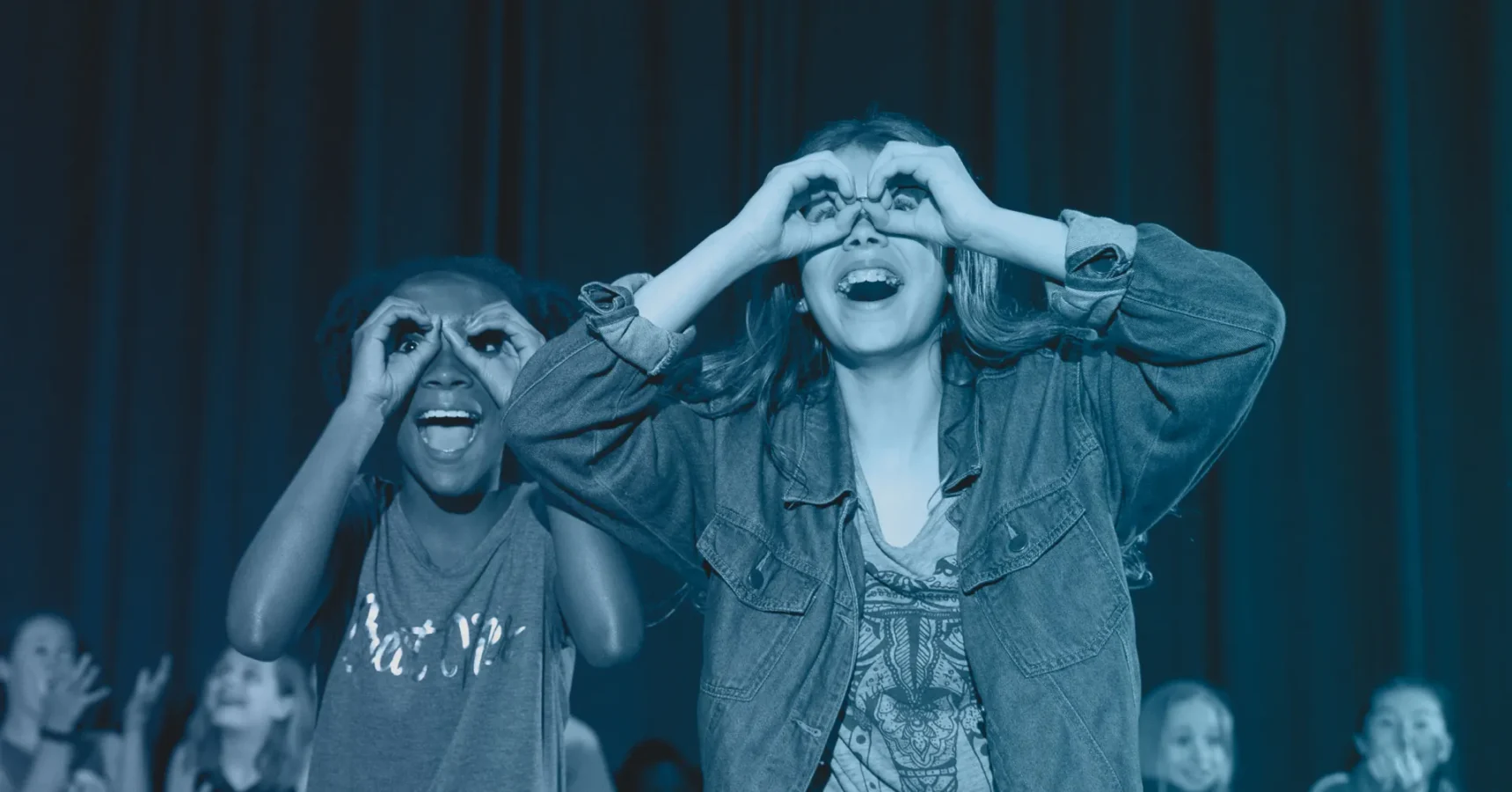 Two children stand side by side, smiling and making pretend binoculars with their hands in front of their faces, with other children visible in the background.